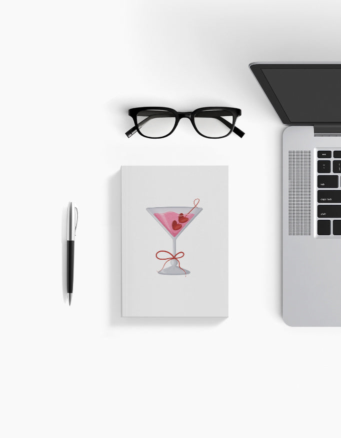 Aesthetic white A5 notebook with pink cocktail glass design – flat lay with glasses, pen, and laptop for work or journaling